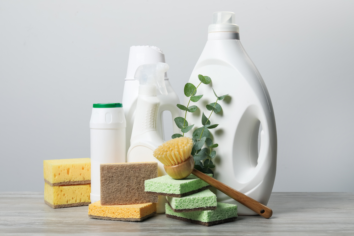 Cleaning supplies including bottles, sponges, and a brush on a wooden surface with a gray background.