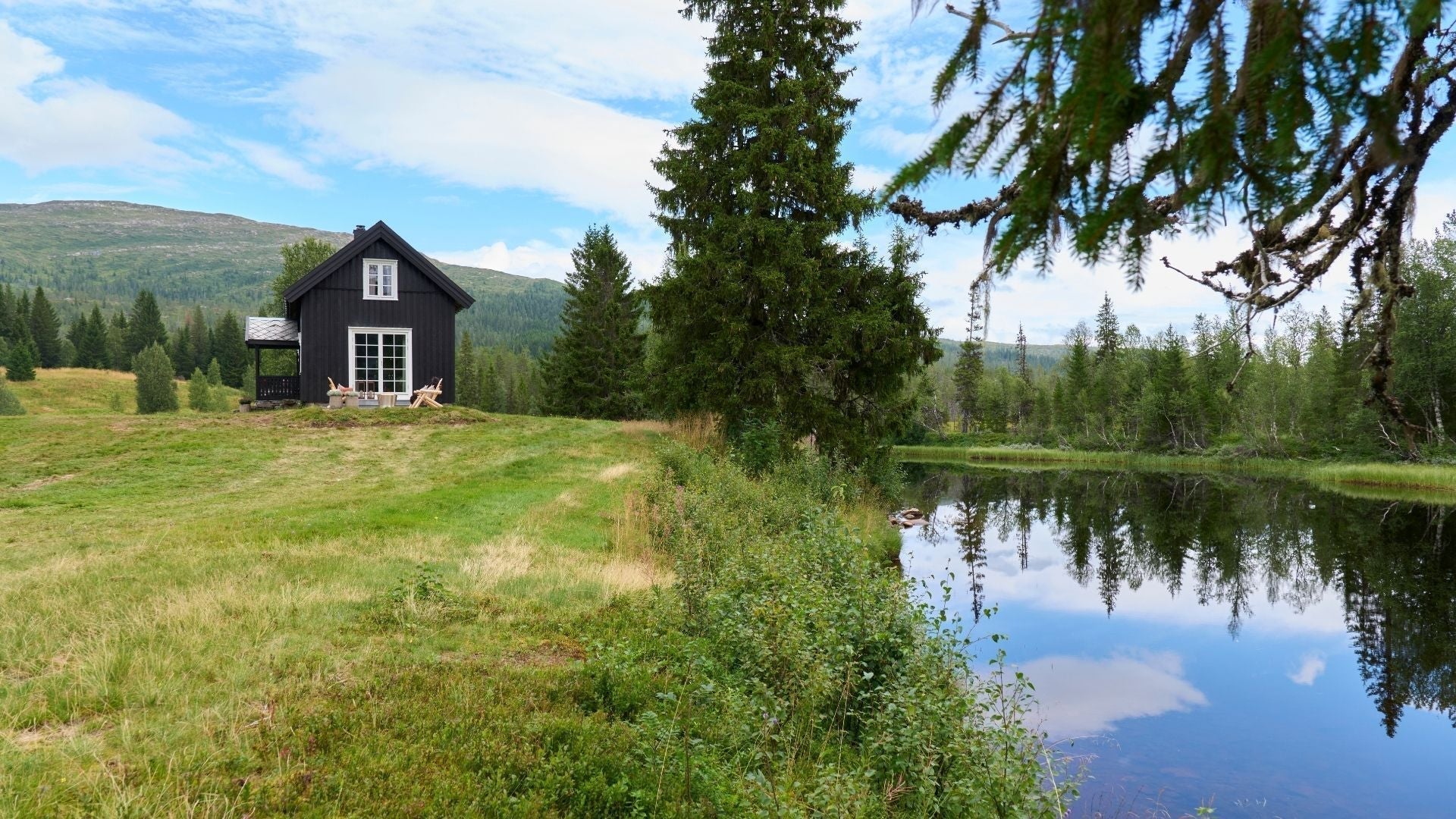 Viihtyisä mökki järven rannalla, taustalla vuoret. 