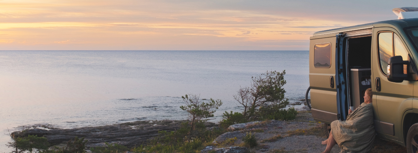 Person sitting in an open van door overlooking a scenic ocean view at sunset.