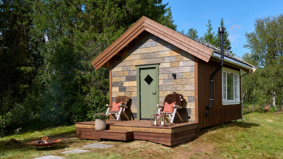 Small wooden cabin in a forest setting with a deck and outdoor furniture.