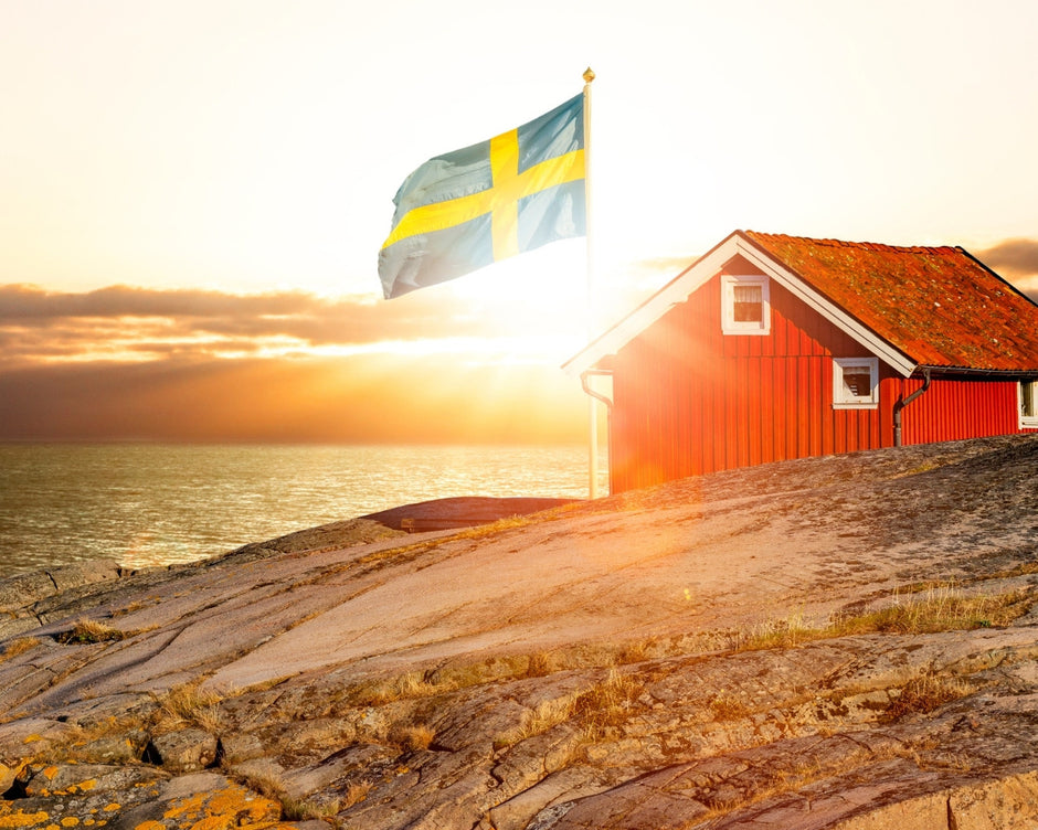 Red cottage with a Swedish flag on a rocky outcrop over water at sunset